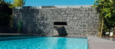 Mauer aus Gabionsteinen mit einem Wasserelement, dass in einen Pool plätschert.
