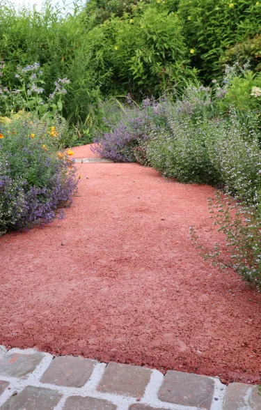 Gehweg aus rotem offenporigem Beton umsäumt von Blumen.