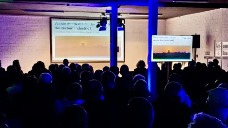 Abgedunkelter Vortragsraum mit Publikum von Hinten und einer Leinwand