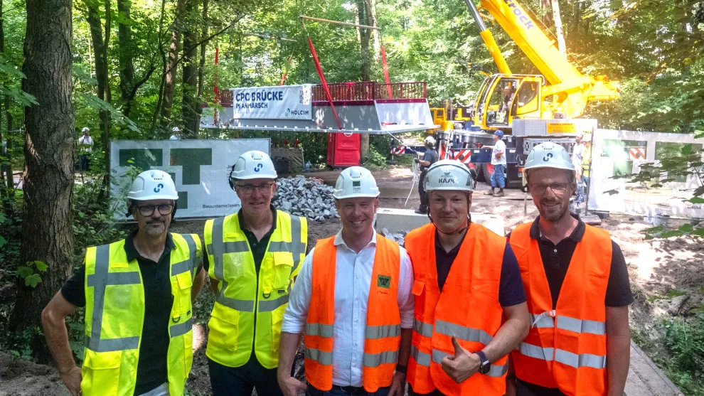 5 Personen in Schutzbekleidung vor einem Kran an dem eine Carbonbetonbbücke hängt.