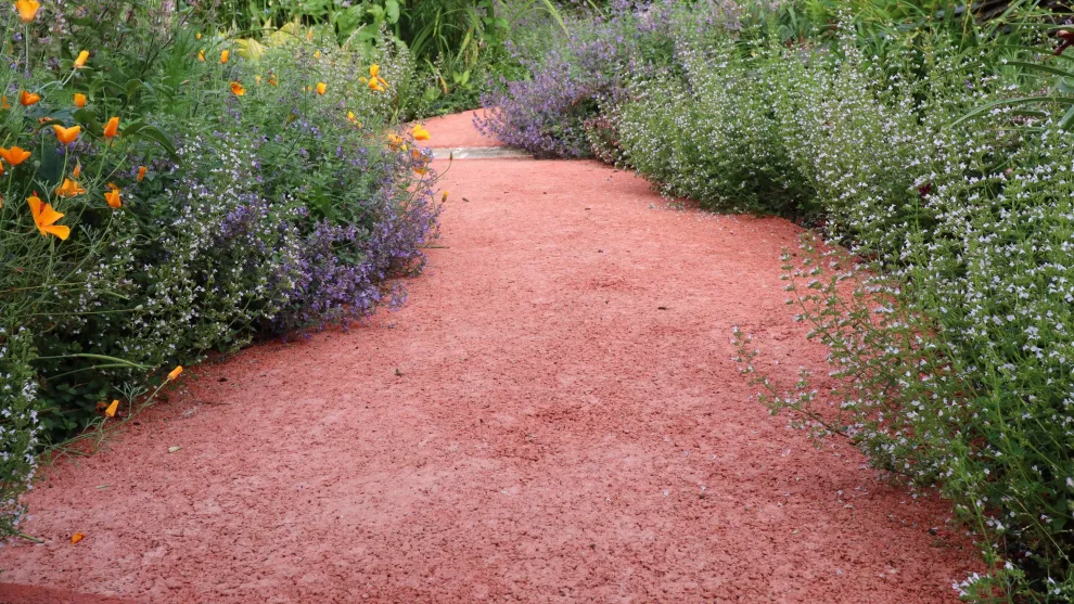 Gehweg aus rotem offenporigem Beton umsäumt von Blumen.