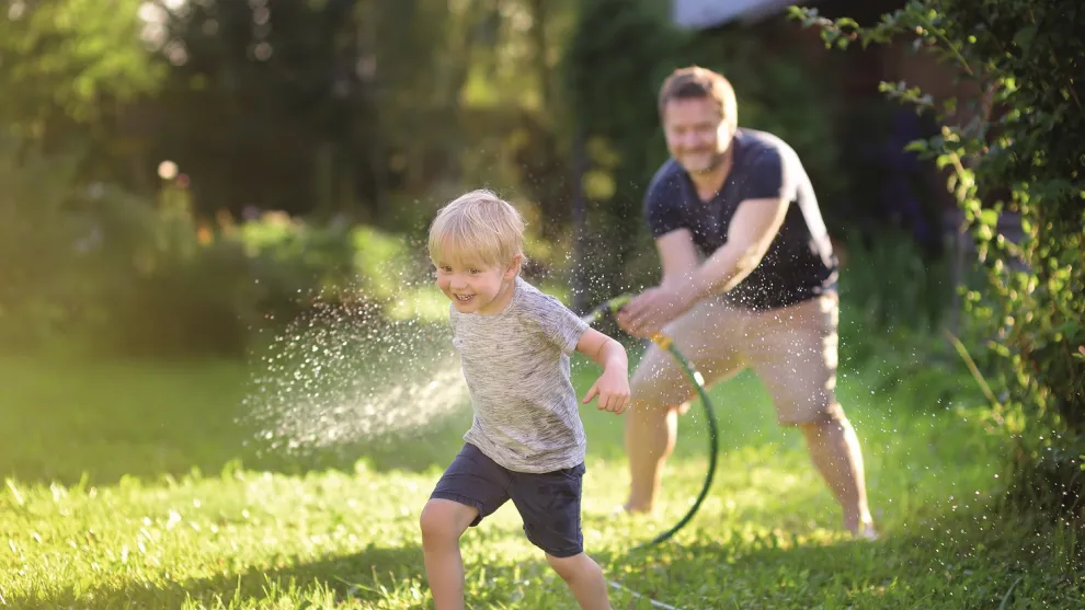 Mann hält einen Gartenschlauch auf ein Kind auf einem Rasen. 