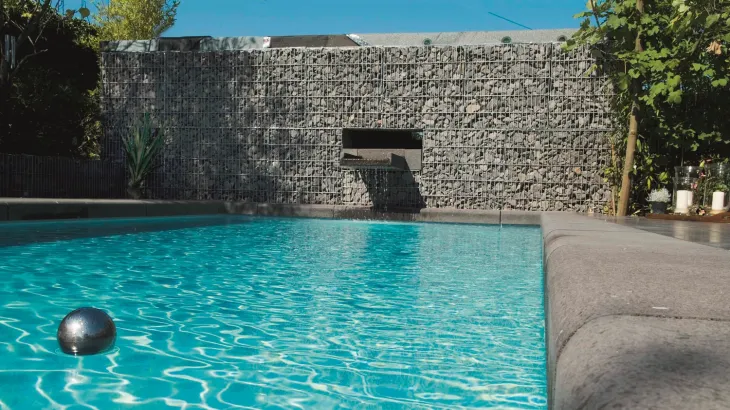 Pool mit einer Wand aus Gabionsteinen aus der ein Wasserzulauf Wasser in den Pool fließen lässt.