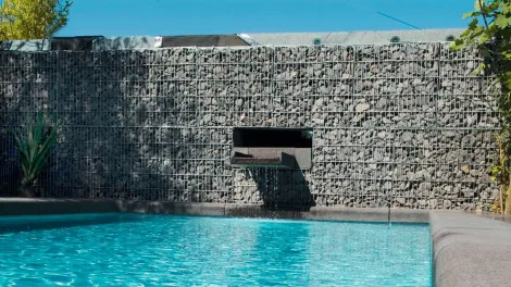 Mauer aus Gabionsteinen mit einem Wasserelement, dass in einen Pool plätschert.